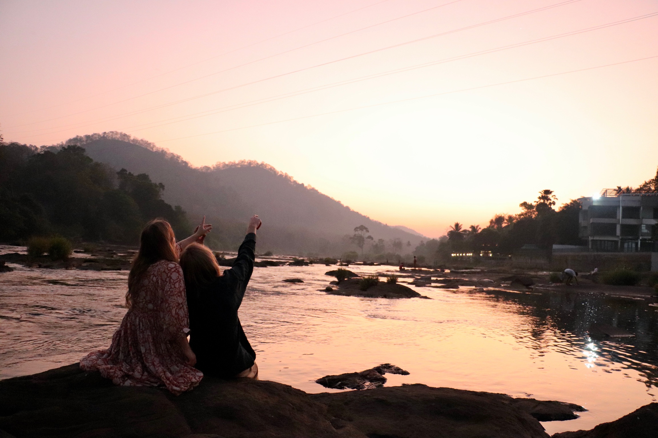 Hanna en haar vriend zitten op een rots bij het water tijdens een zonsondergang. De lucht is een mix geel- en oranjetinten. Hanna en haar vriend wijzen naar de mooie lucht.
