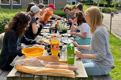 Meet & eat groep. Tien jongeren zitten buiten aan een lange tafel. Er staat eten en drinken op de tafel.