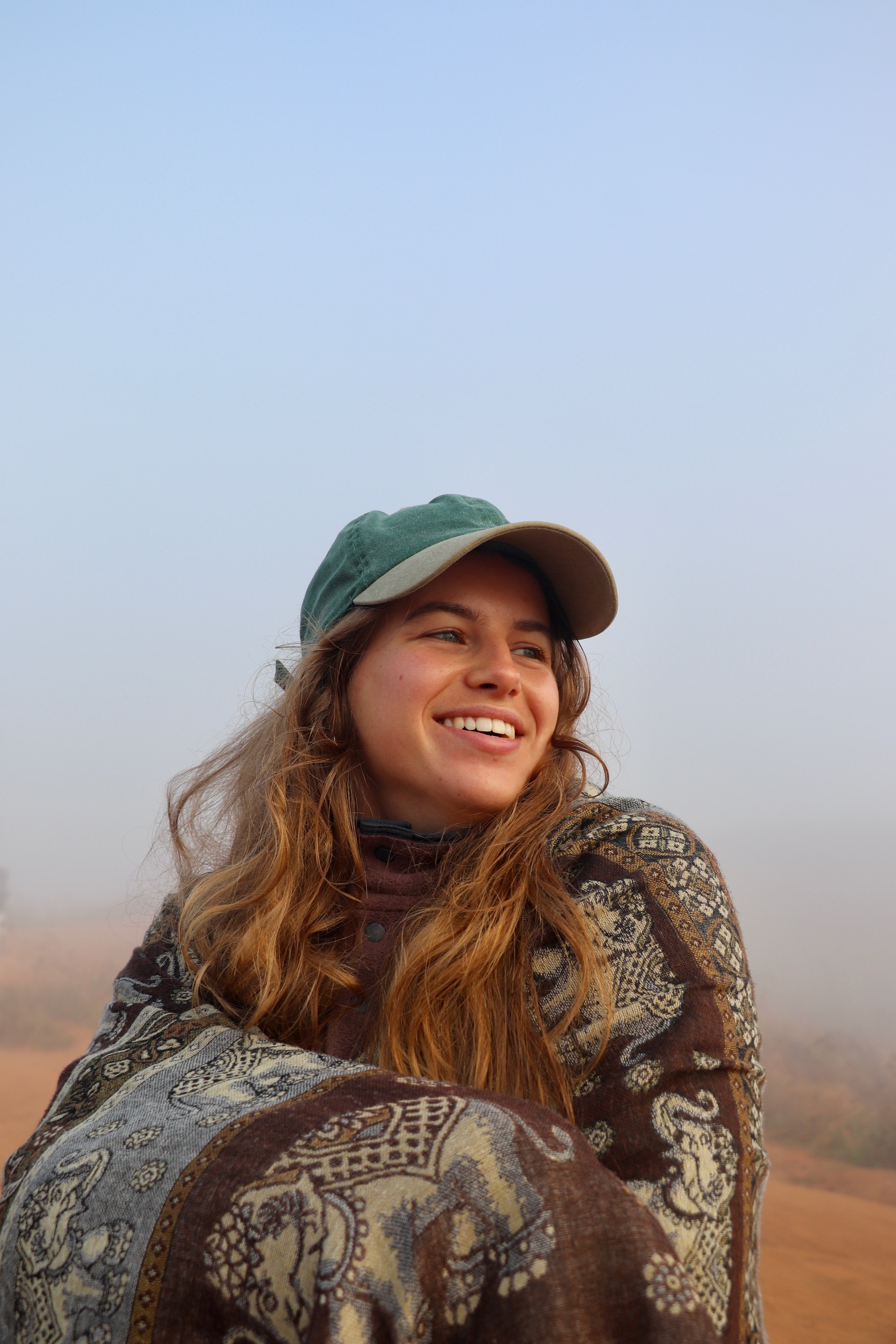 Een close-up van Hanna. Ze zit in een zanderige omgeving. Ze draagt een pet, en kijkt opzij, weg van de camera. Ze heeft een blije, tevreden blik op haar gezicht.