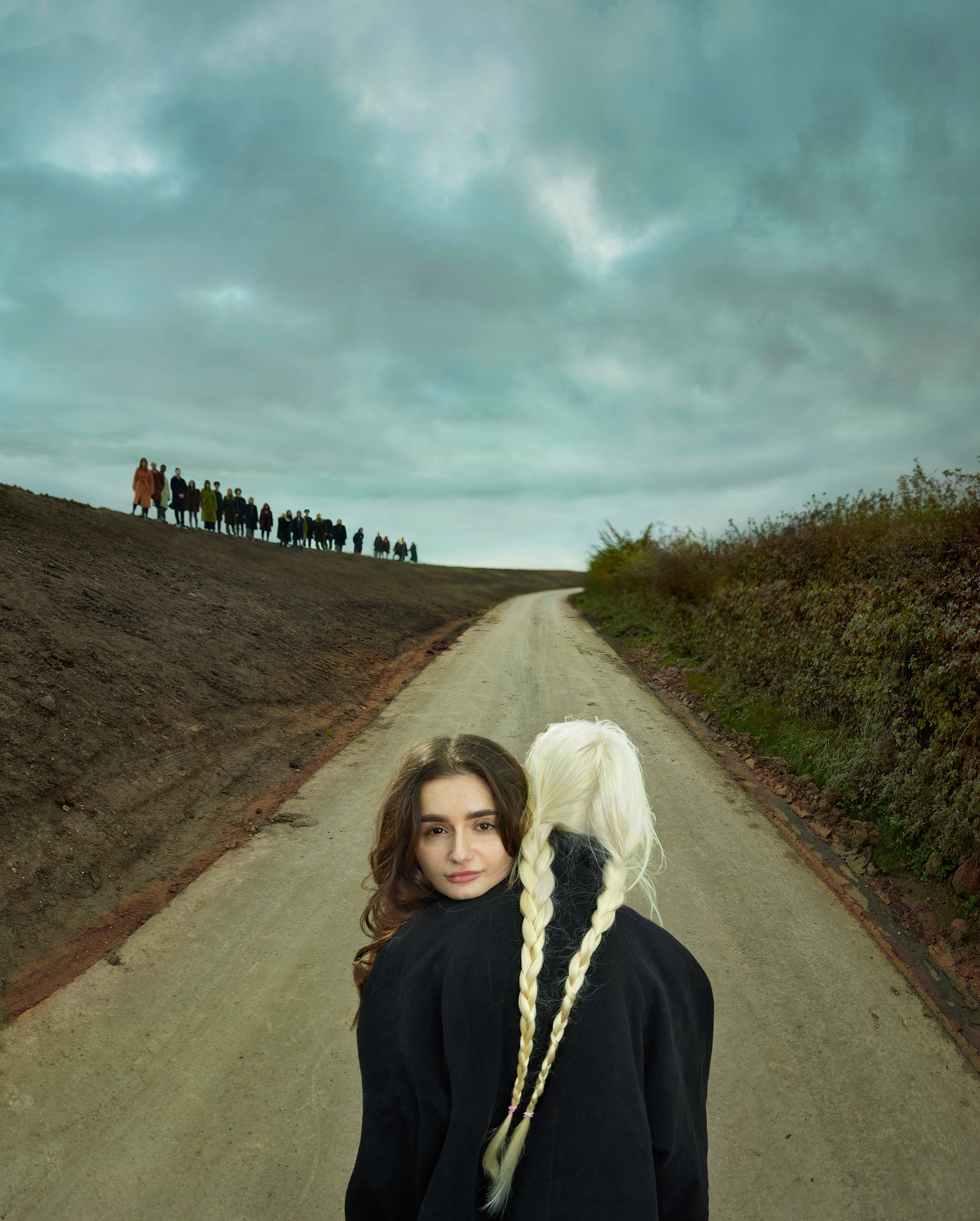 Rana (links) kijkt de camera in. Haar hoofd rust op de schouder van een blond meisje, die met haar rug naar de camera toestaan. Het lichte haar van het meisje en het donkere haar van Rana contrasteren elkaar. Ze staan op een zandpad, met aan de linkerkant een zand-heuvel. Hier staat een rij, gemaakt door de andere acteurs, op. Rechts van het pad is een grassige heuvel.
