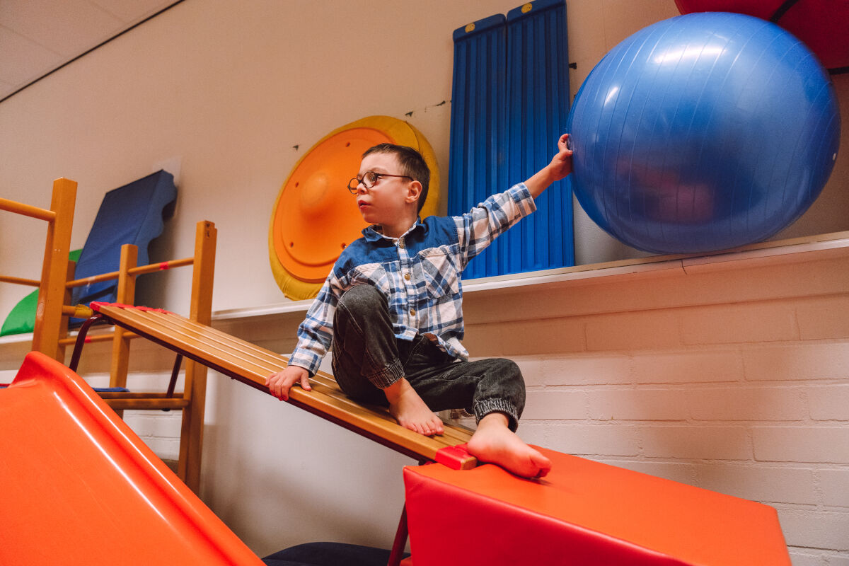 jongen zit op houten bank in gymzaal Visio Amsterdam en houdt een grote blauwe fitnessbal vast