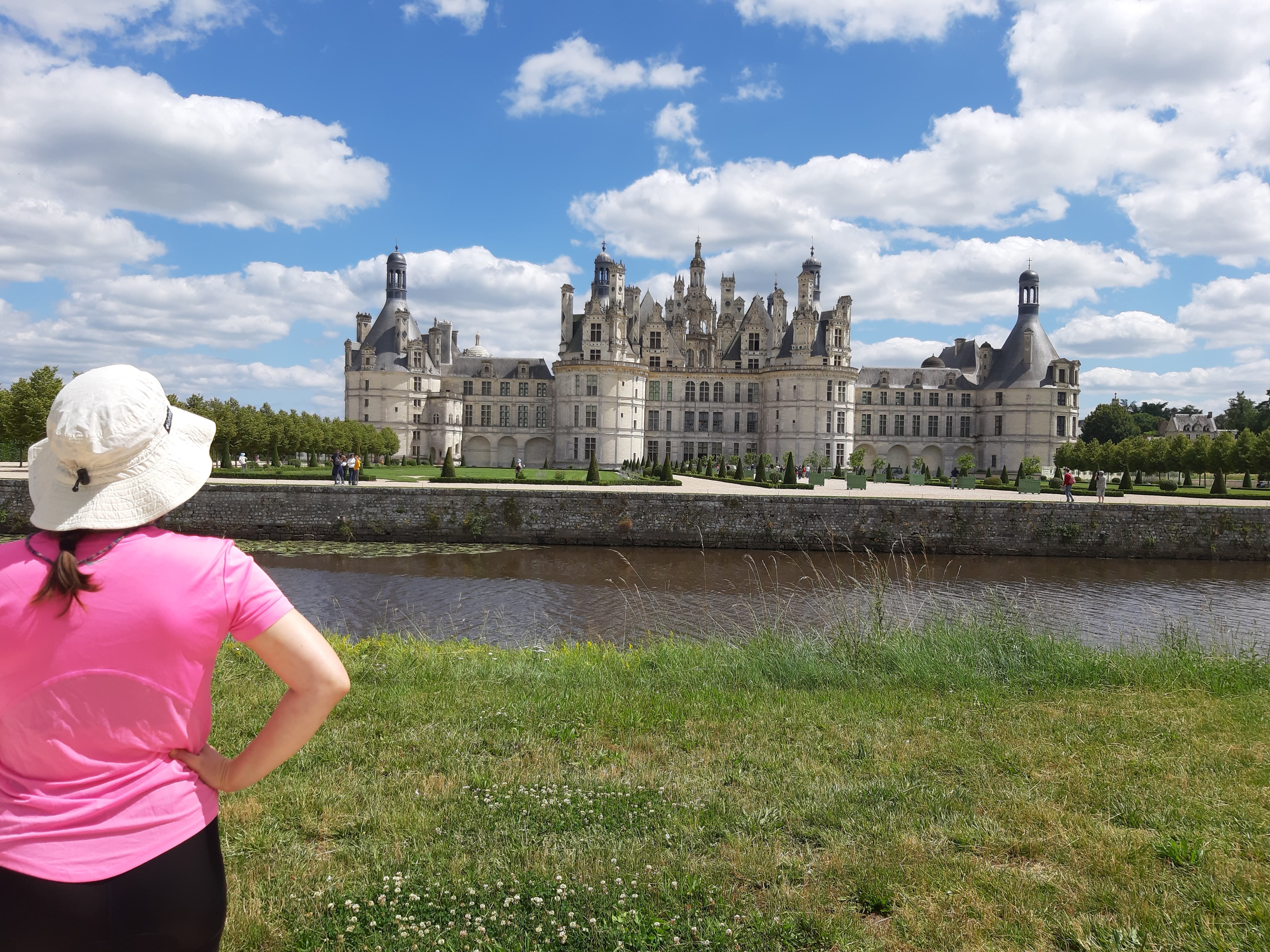 Een vrouw met een roze shirt en een witte hoed kijkt naar een kasteel aan de overkant van het water.
