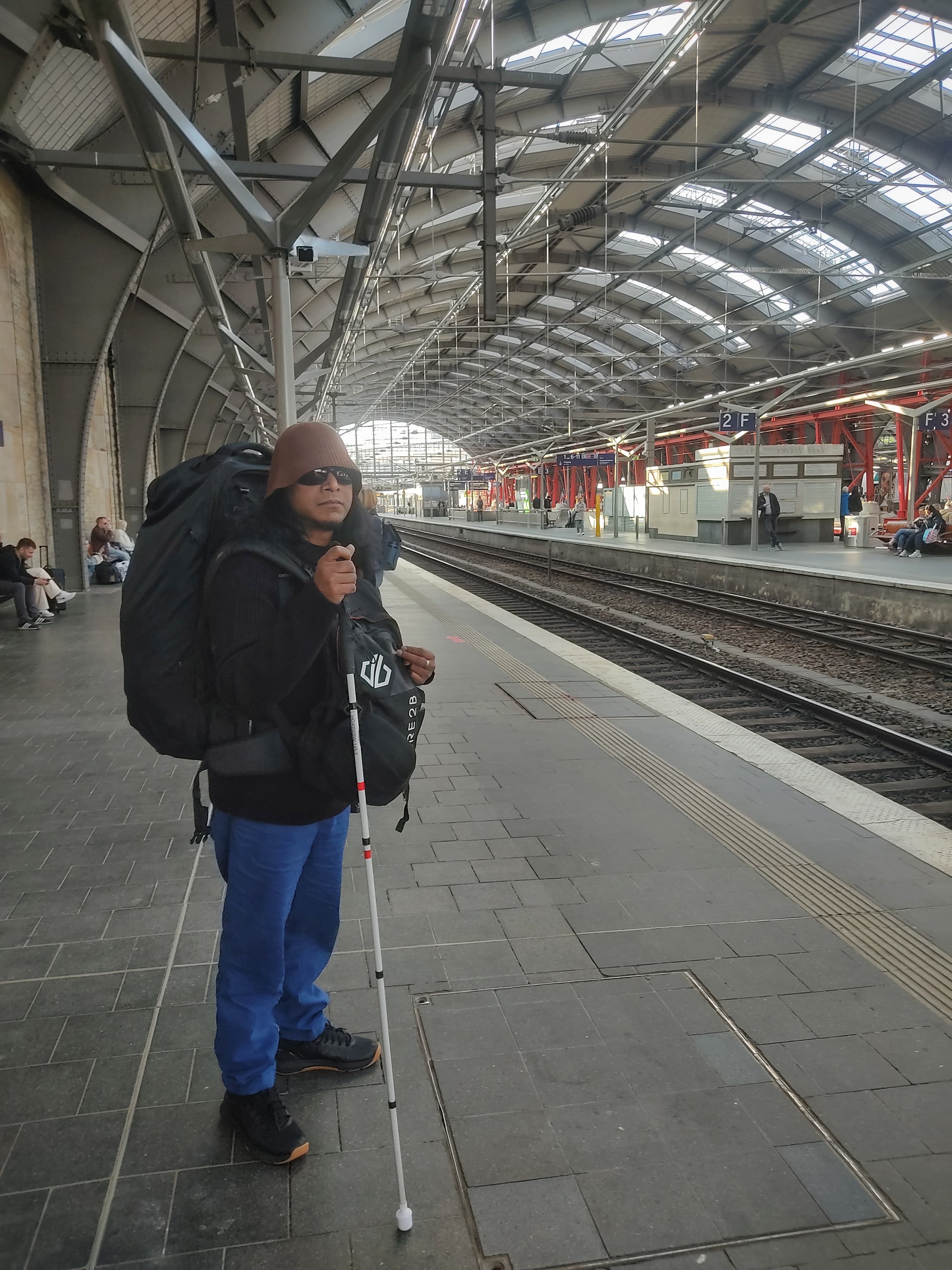 Emmanuel staat op een perron te wachten op een trein. Hij heeft lang zwart haar en een baardje, en draagt een zonnebril en een muts. Hij heeft een grote rugzak op zijn rug, en een kleine tas voorop. Hij heeft zijn taststok vast.