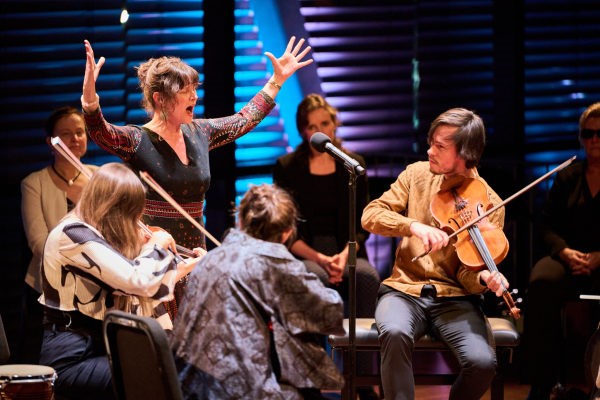 Je ziet een vrouw zingen met grote gebaren. Daarnaast drie mensen die op strijkinstrumenten spelen.