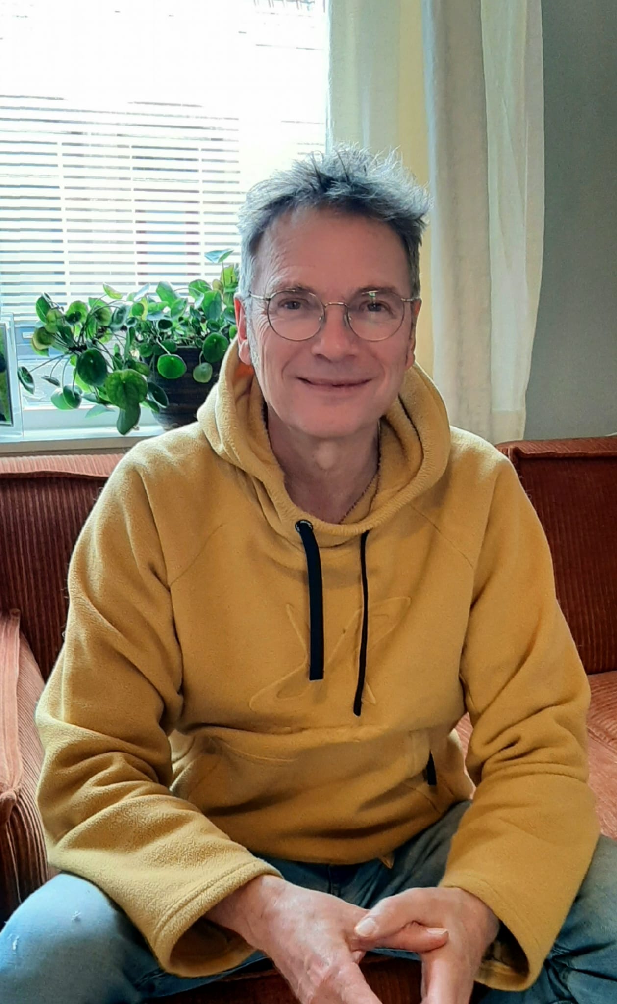 Chris heeft een bril op, zit met een gele trui aan op de bank.