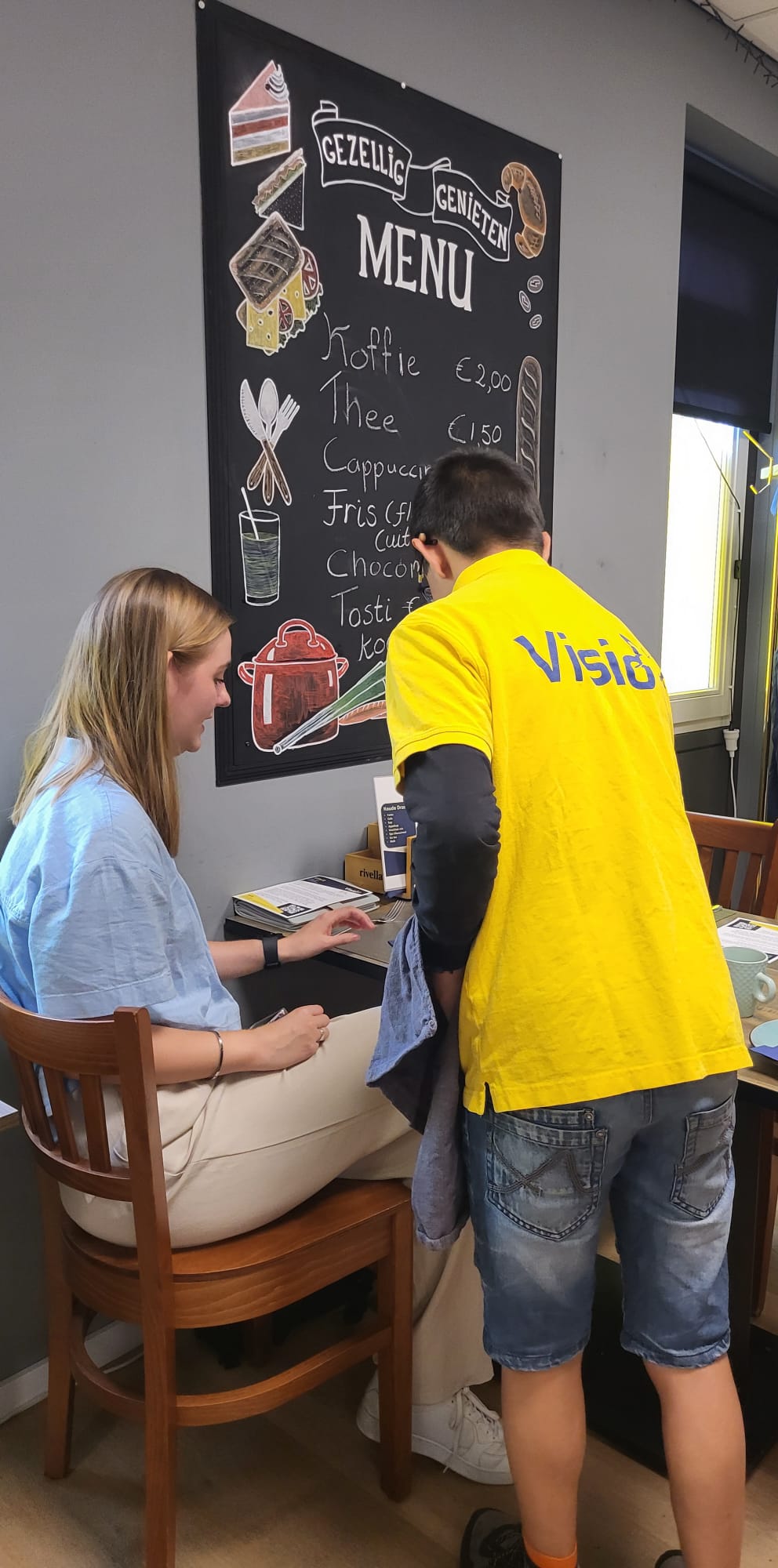 Ivan serveert aan een tafeltje. Hij heeft donker haar, en draagt een geel shirt met Visio erop.