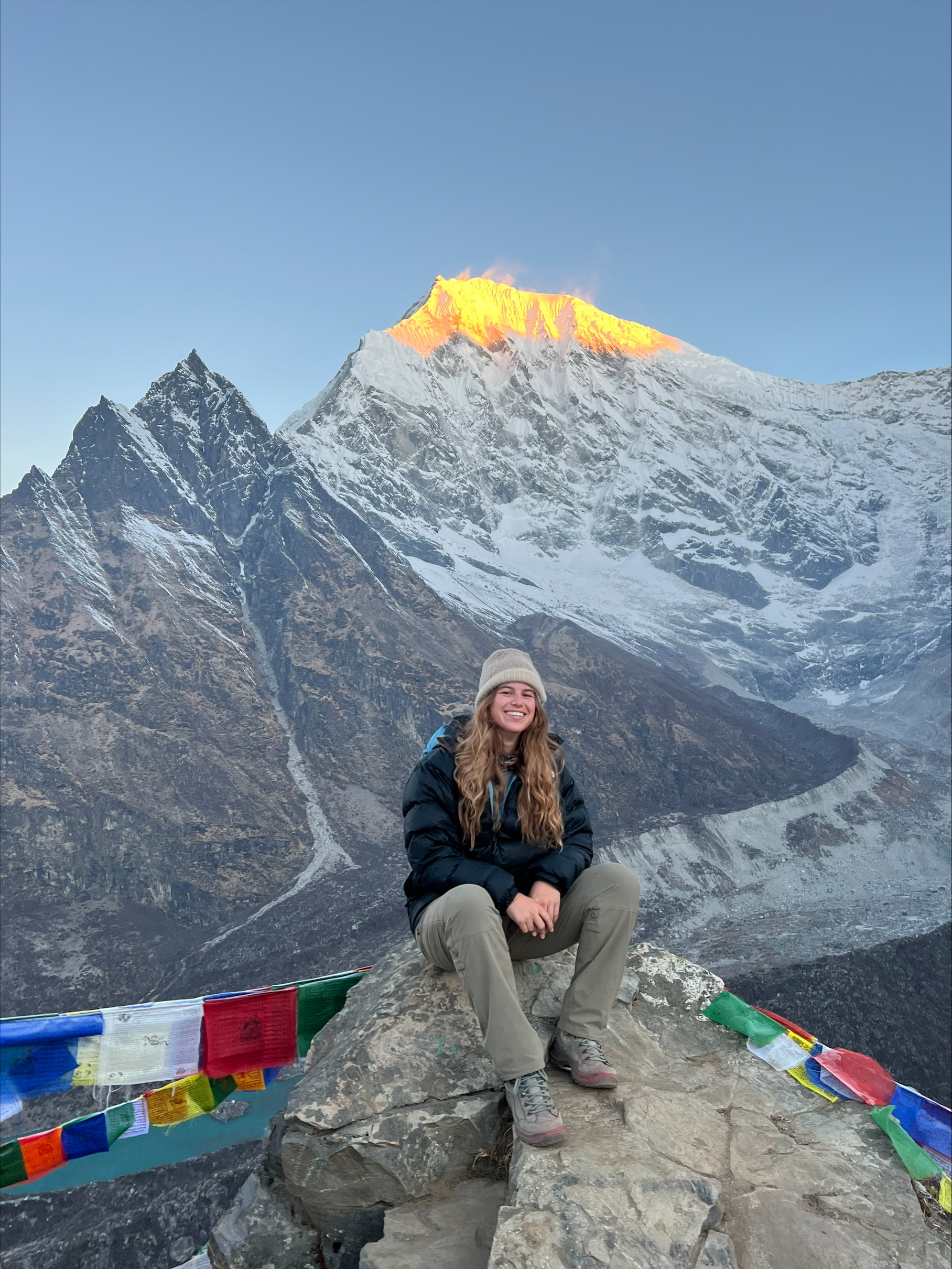 Hanna zit op een rots. In de achtergrond zijn hoge bergen te zien. De zon schijnt op de top van de hoogste berg, waardoor er een warme, oranje gloed te zien is. De foto is genomen in Nepal.
