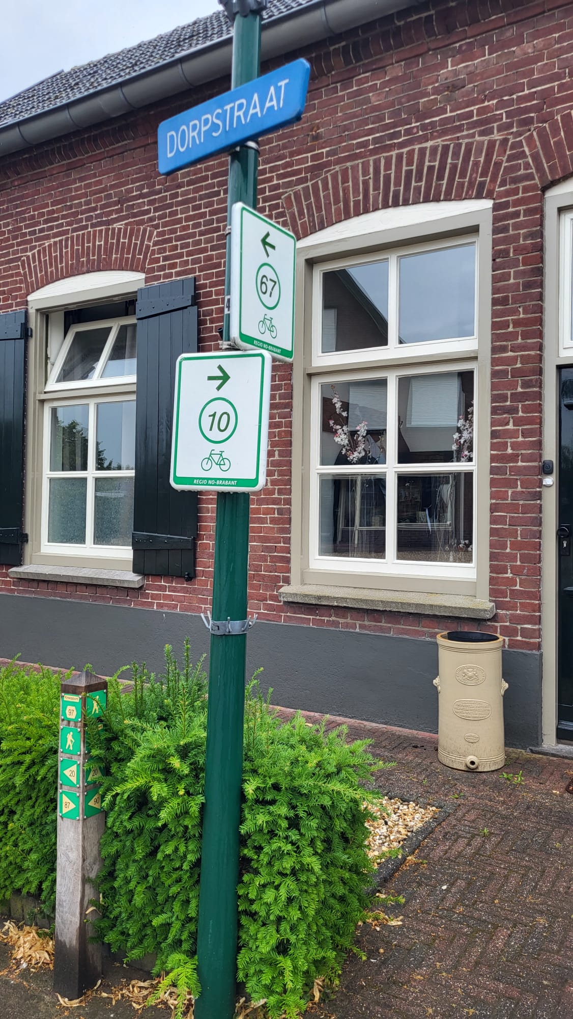 Een gebouw is te zien op de achtergrond. Op de voorgrond is een lantaarnpaal met een blauw straatnaambordje met "dorpstraat" erop. Op de paal zijn ook twee groen/witte bordjes van fietsroutes te zien. De bovenste wijst naar links en heeft het nummer 67, de onderste wijst naar rechts en heeft het nummer 10.