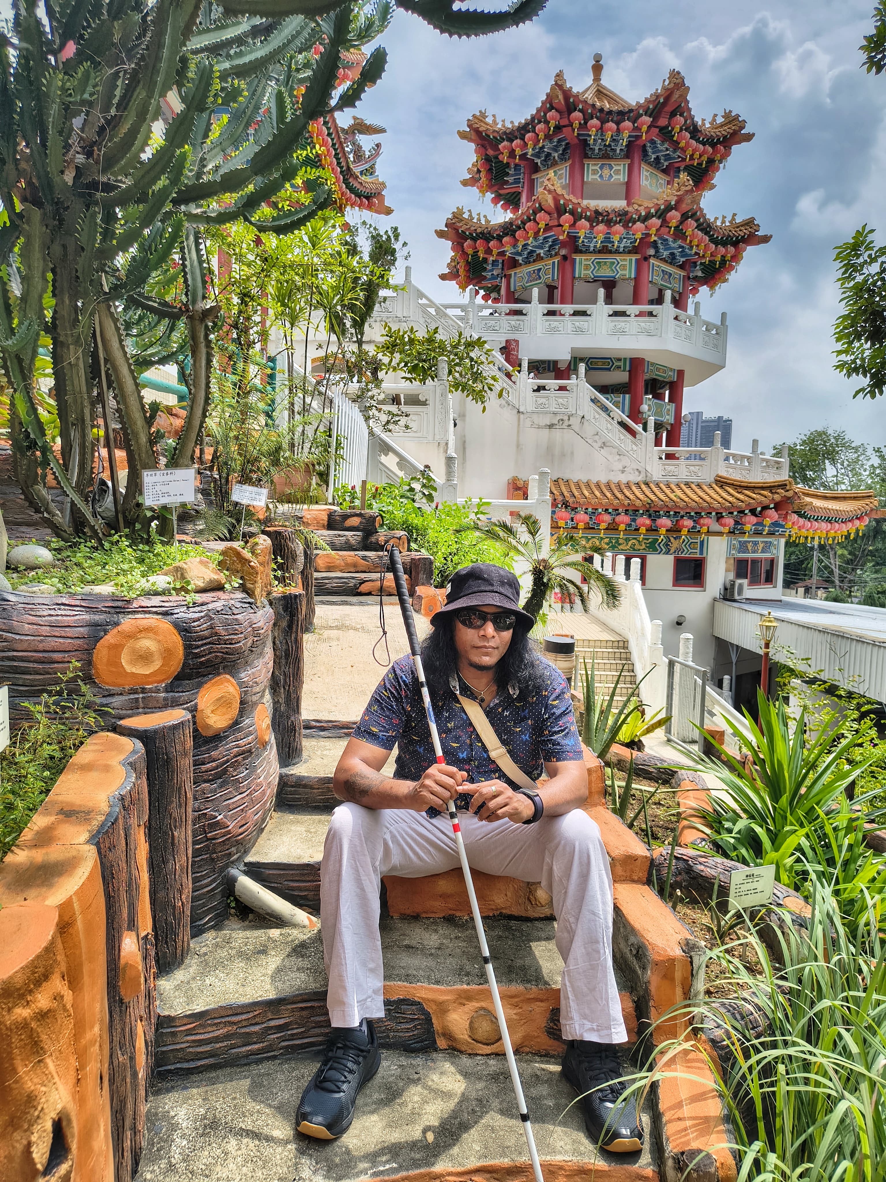 Emmanuel zit op een trap, met zijn taststok vast. Op de achtergrond is een mooie kleurrijke toren te zien.