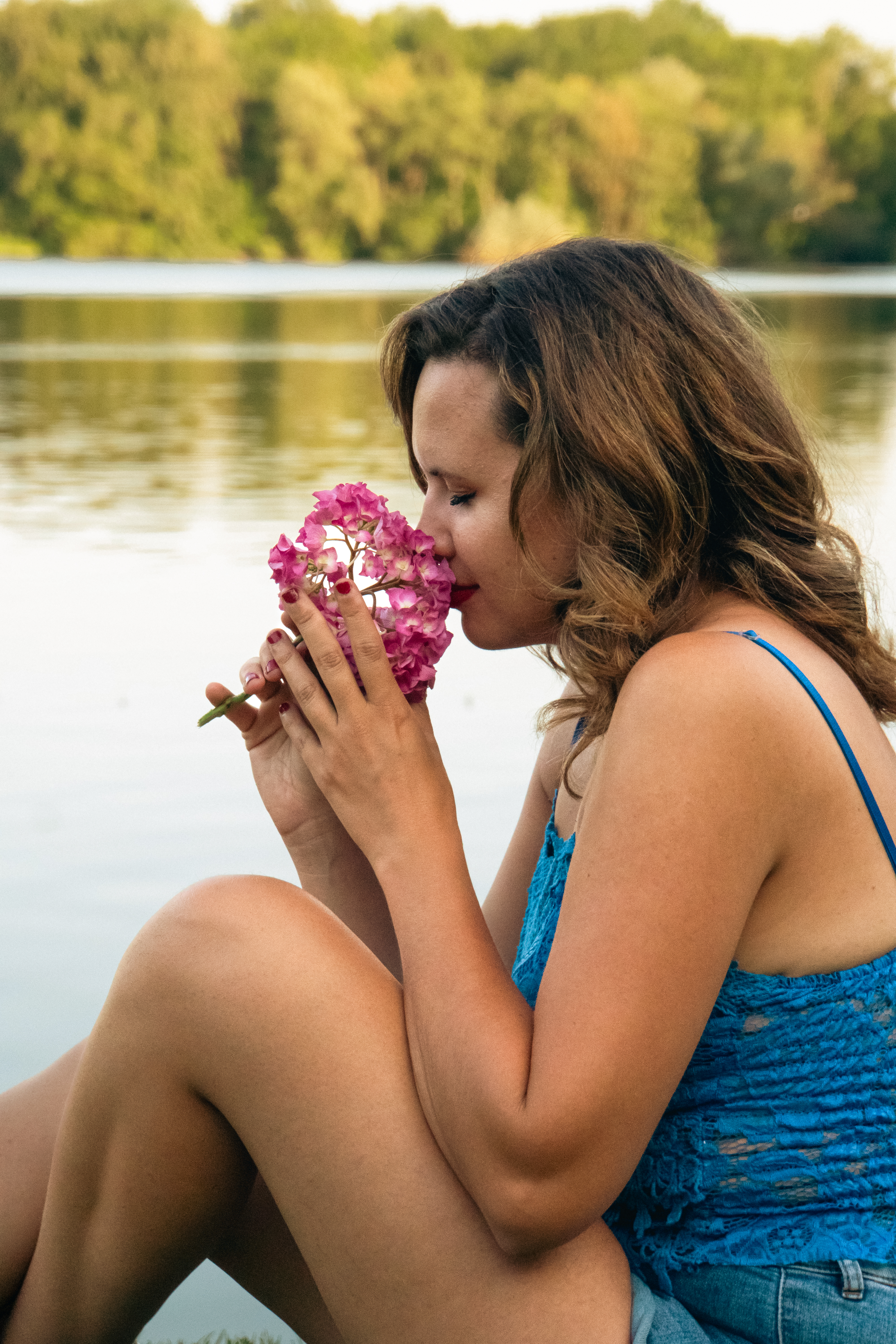 Marie Elise zit aan het water, in de zon. Ze heeft zomerse kleding aan, en ruikt aan een felroze bloem.