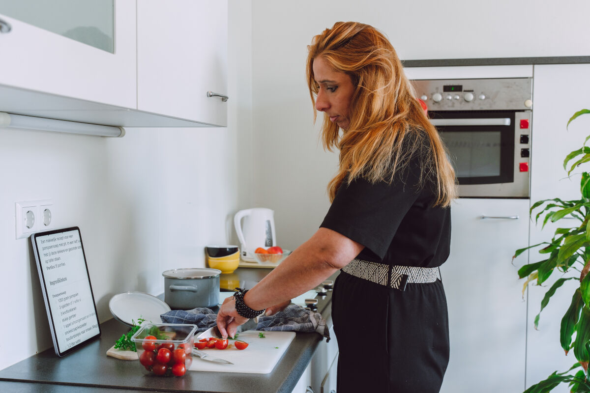 Dame is aan het koken en snijdt tomaatjes op een snijplank en volgt een vergroot recept op een tablet