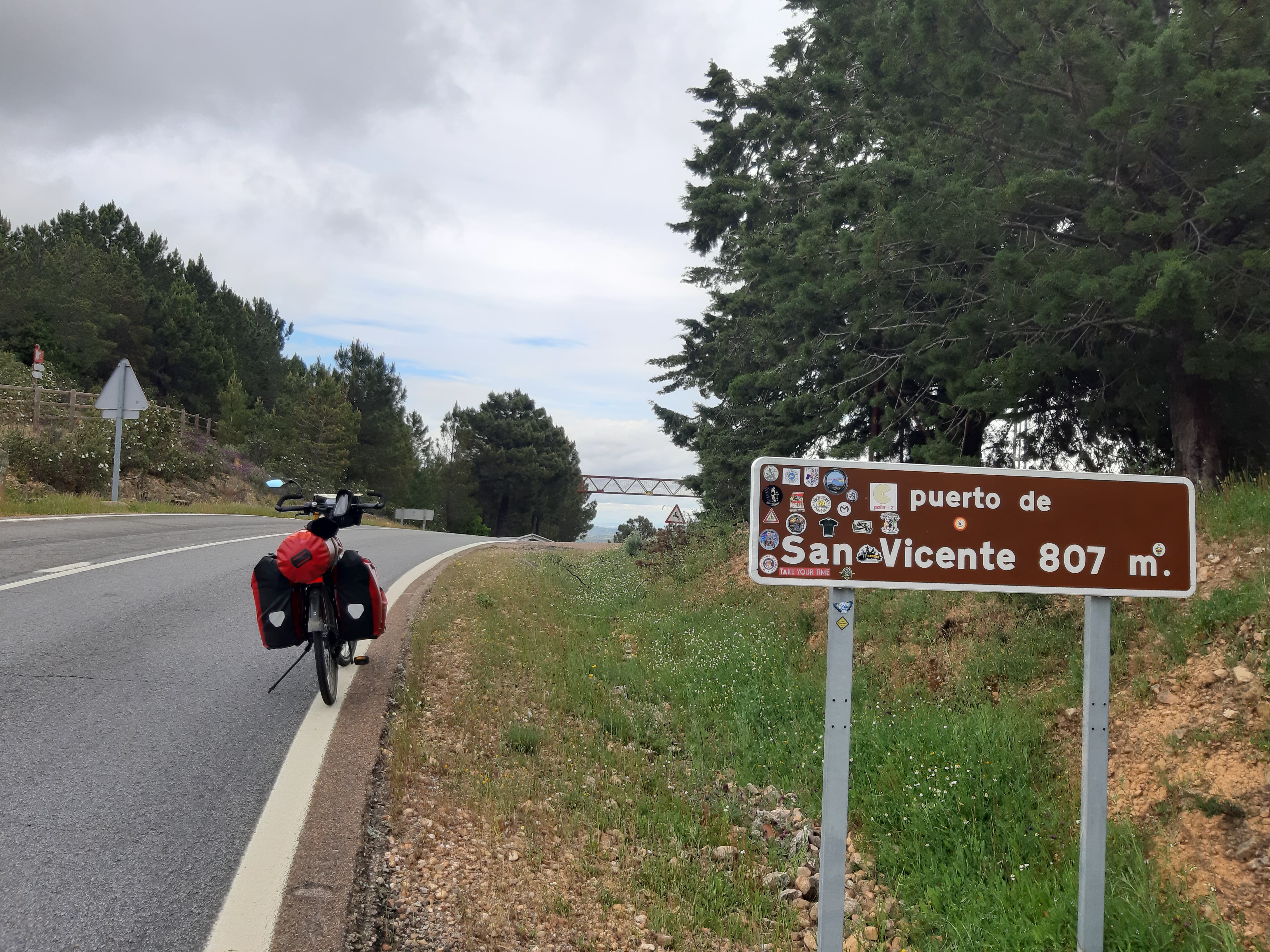 De fiets van Lex, met een rode tas achterop, staat op de weg. Naast de weg staat een bord met "puertde de Vicente 807" erop.
